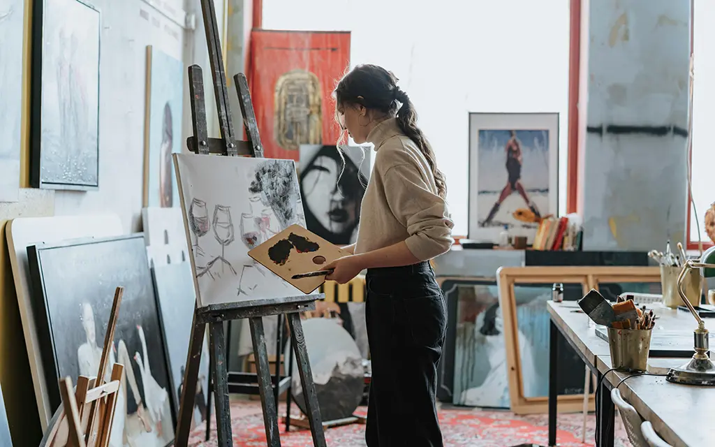 A young artist painting on a canvas in a bright studio, surrounded by artworks, sketches, and painting tools
